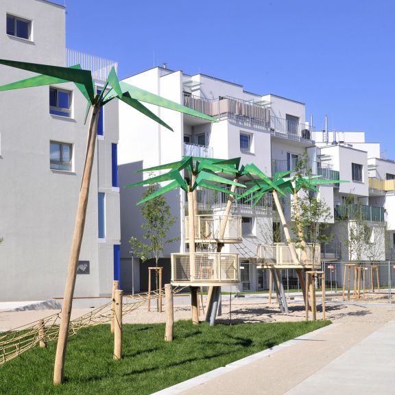 Hellgraue Fassade mit verschiedengroßen Fenster sowie Balkonen Begrünung mit Holz Metall Palmen für die Kinder © Gesiba