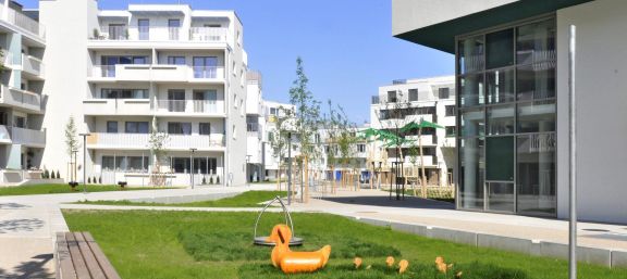 Weißes Eckiges Gebäude mit Balkonen hofseitig viel Freifläche für Kinder mit Spielplätzen schön grün. © Gesiba